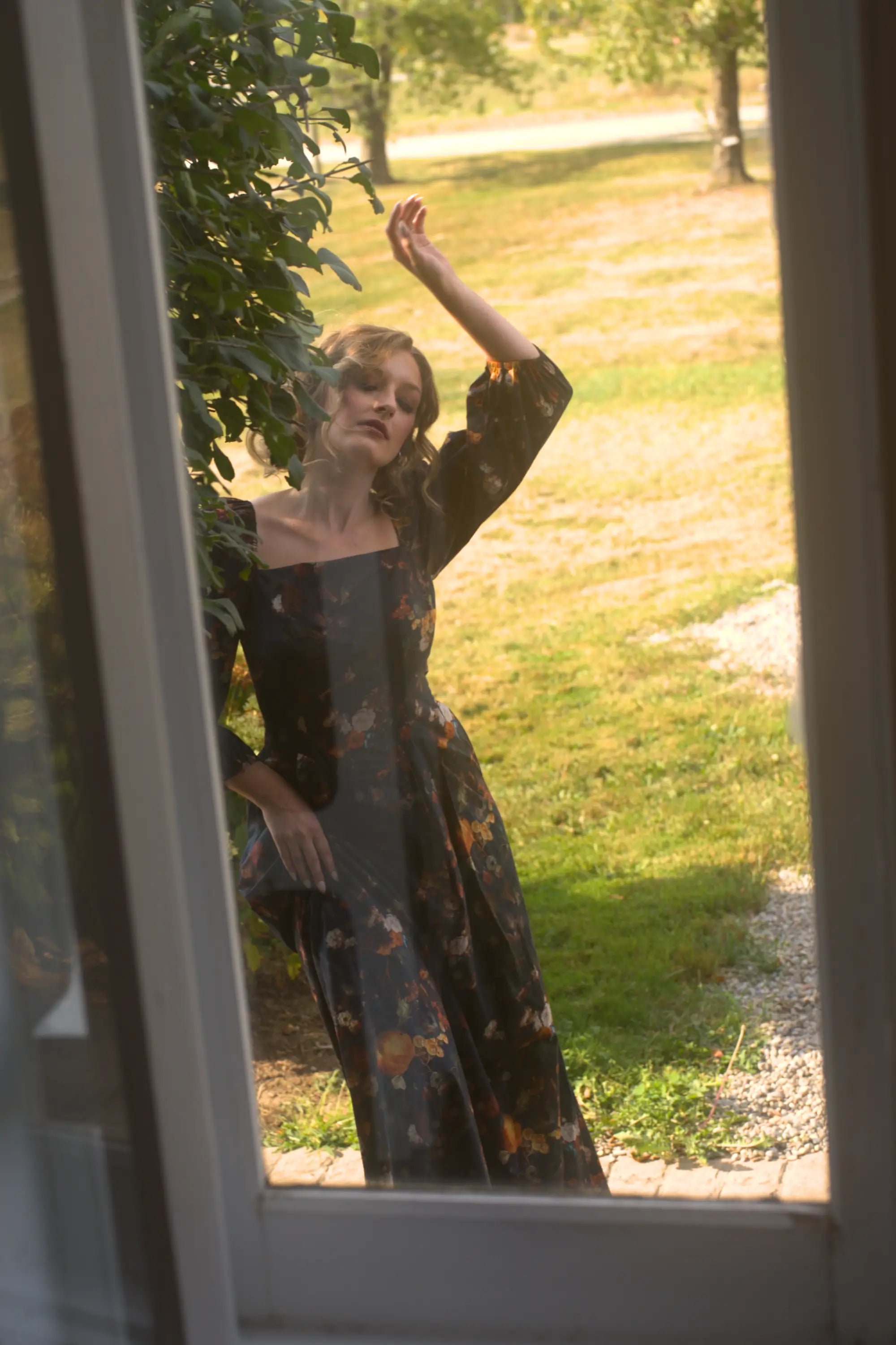 A woman in a floral cotton maxi dress standing outdoors, viewed through a glass door.