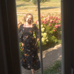 A woman in a floral cotton maxi dress standing in a garden, viewed through a window.