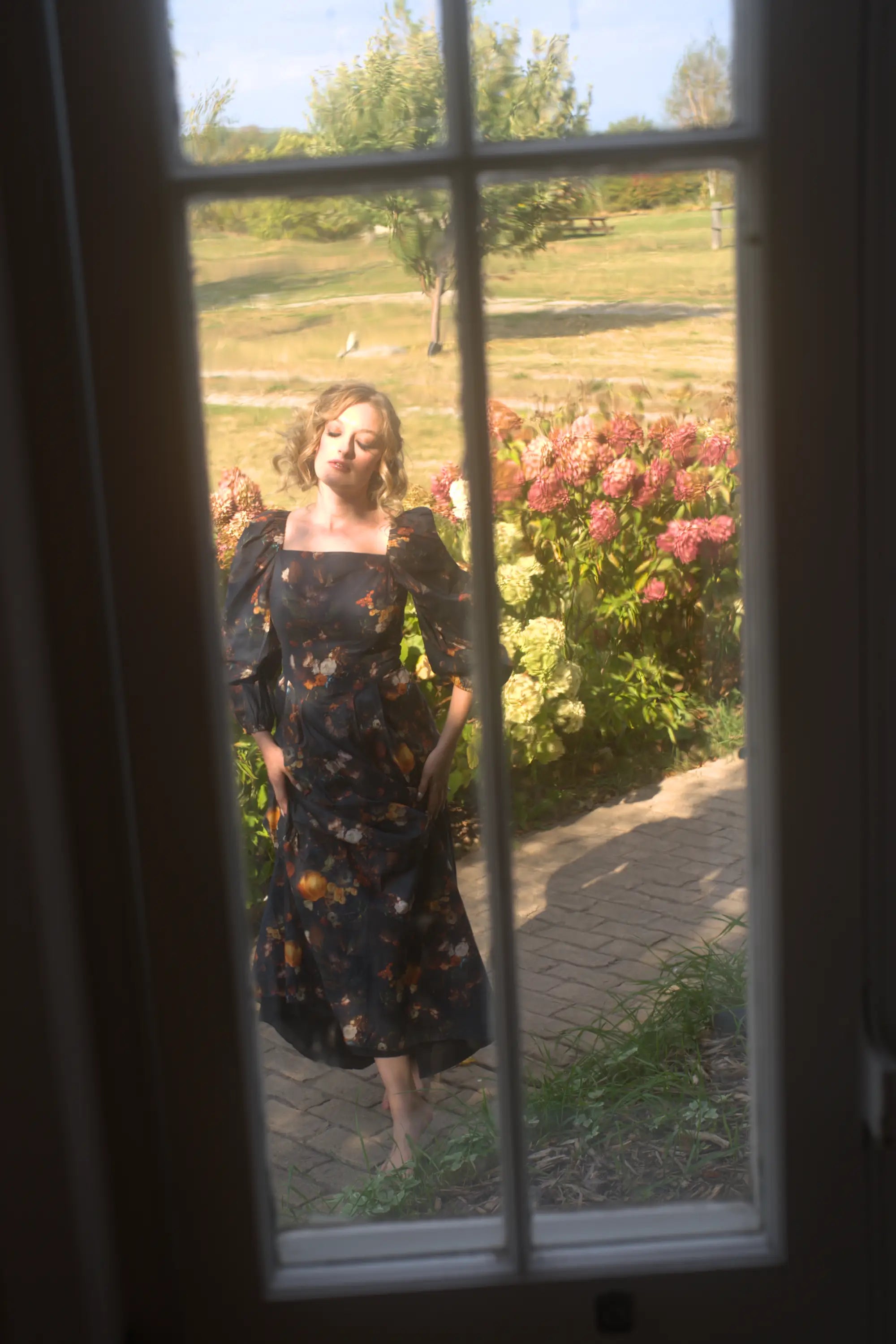 A woman in a floral cotton maxi dress standing in a garden, viewed through a window.