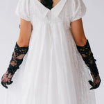 A person wearing a white lace dress with black opera gloves on a plain background