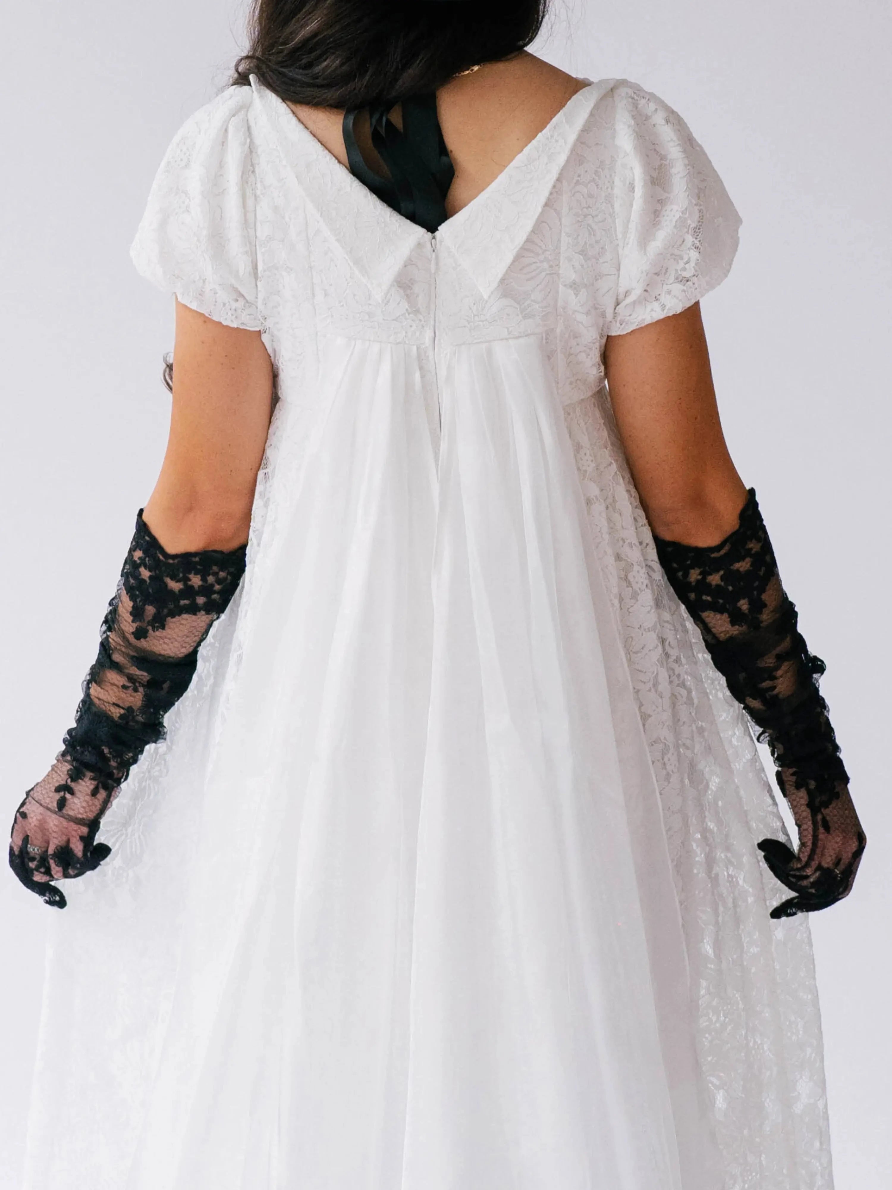 A person wearing a white lace dress with black opera gloves on a plain background