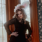 A woman in a black cotton dress with a decorative hat in a room with floral wallpaper.