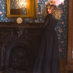 A woman in a black cotton maxi dress is standing in a room with floral wallpaper and a fireplace.