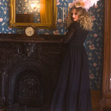 A woman in a black cotton maxi dress is standing in a room with floral wallpaper and a fireplace.