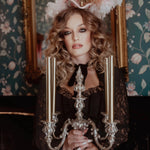 A woman in a black cotton dress holding a silver candelabra in front of a decorative mirror with floral wallpaper.