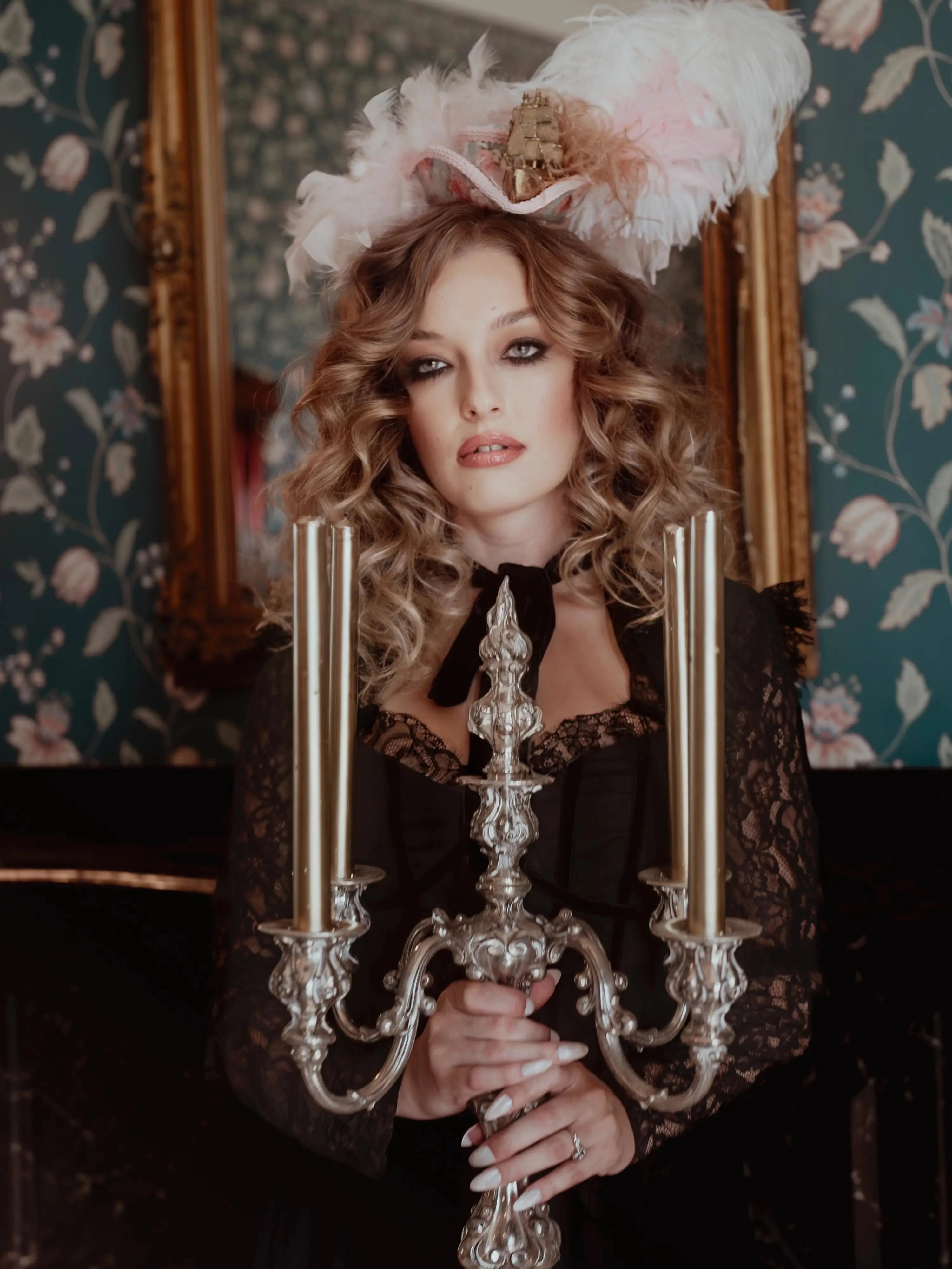 A woman in a black cotton dress holding a silver candelabra in front of a decorative mirror with floral wallpaper.