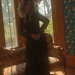 A person in a black cotton lace dress with an antique headpiece is standing in a room with large windows and patterned wallpaper.