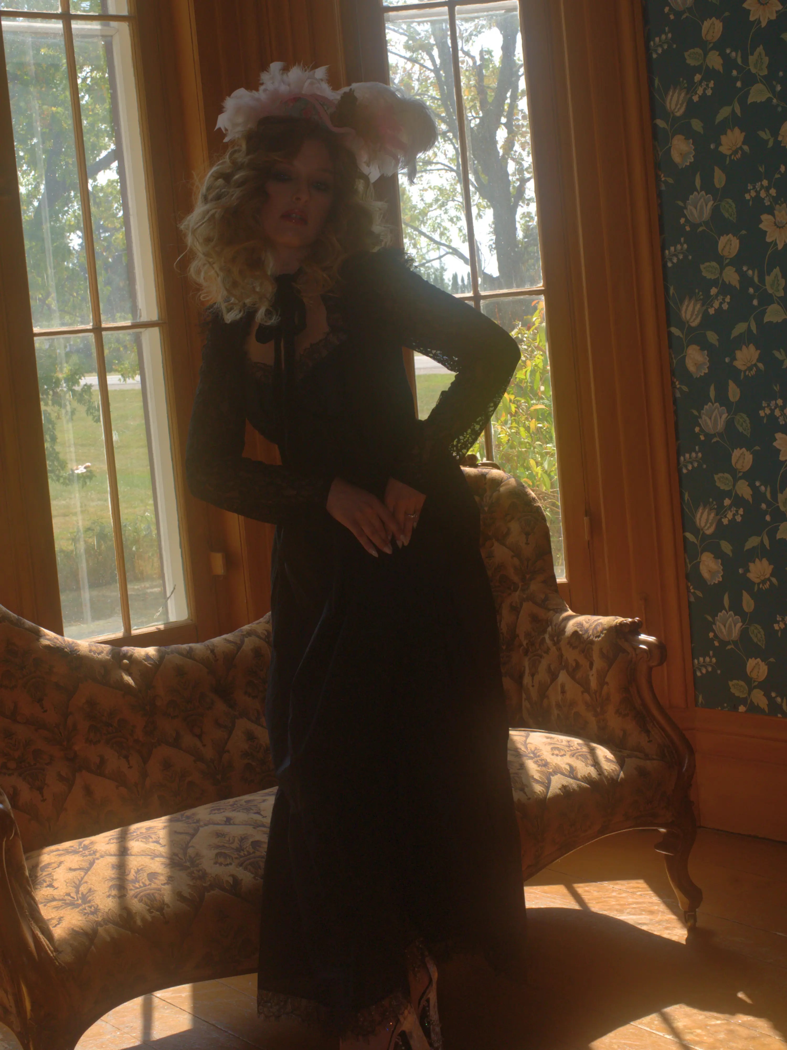 A person in a black cotton lace dress with an antique headpiece is standing in a room with large windows and patterned wallpaper.