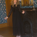A person in a black Eveline lace dress is standing in a room with floral wallpaper and a fireplace.