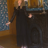 A person in a black Eveline lace dress is standing in a room with floral wallpaper and a fireplace.