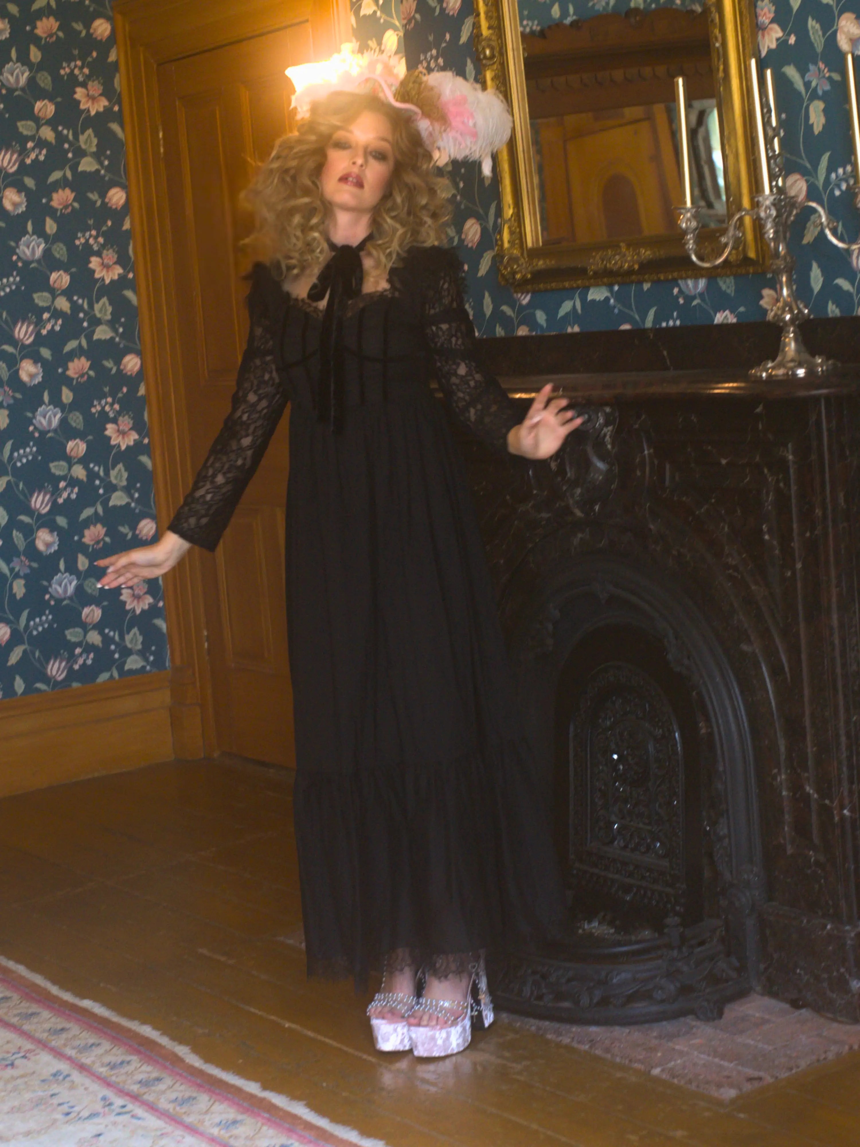 A person in a black Eveline lace dress is standing in a room with floral wallpaper and a fireplace.