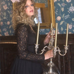 A woman in a black long lace sleeve dress holding a silver candelabrum against a decorative wall.