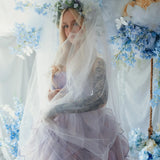 A woman wearing a lavender tulip dress and a white tulle veil surrounded by floral arrangements.