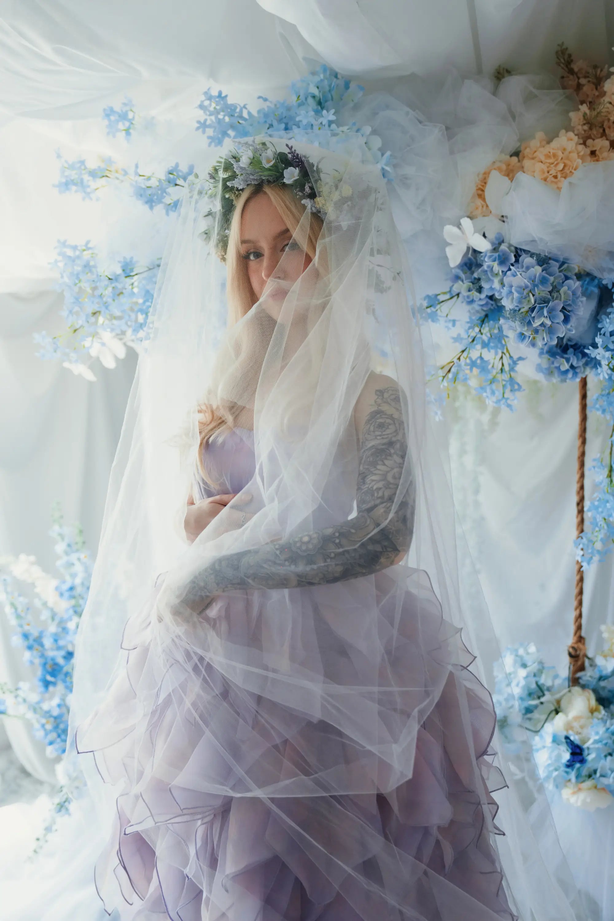 A woman wearing a lavender tulip dress and a white tulle veil surrounded by floral arrangements.