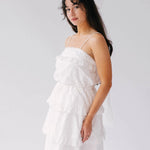A woman wearing a white dress with a bridal hair bow in her hair, against a plain background.
