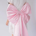 A woman wearing a pink bow belt draped over a white lace dress on a plain background.