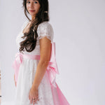A woman wearing a white lace dress with the Ballet Pink Grand Bow Belt for a stylish look.