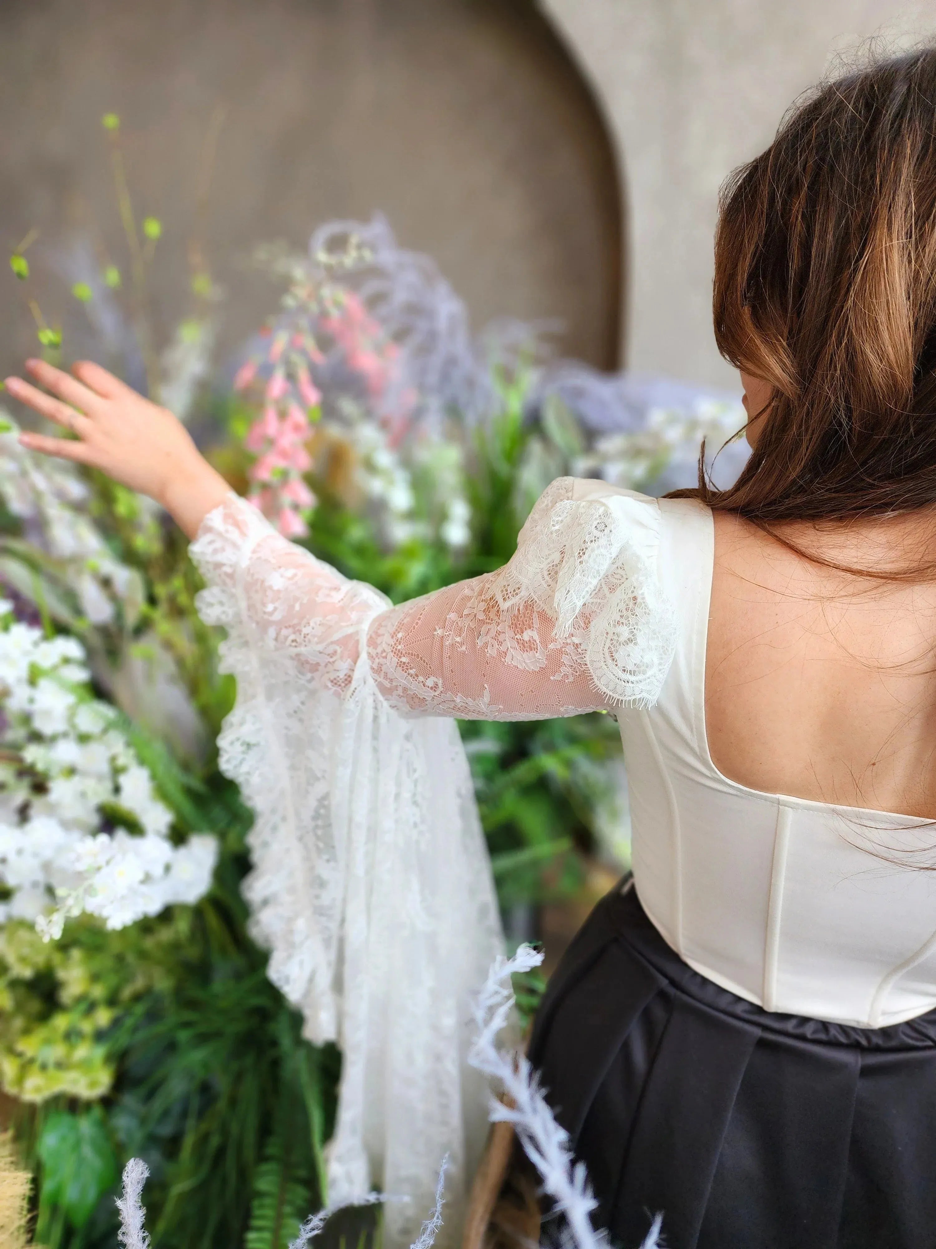 A woman in a white lace corset top, showcasing the billowing lace sleeves, surrounded by florals.