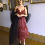 A woman wearing a red organza midi dress with a black tulle giant bow sash in an art gallery setting.