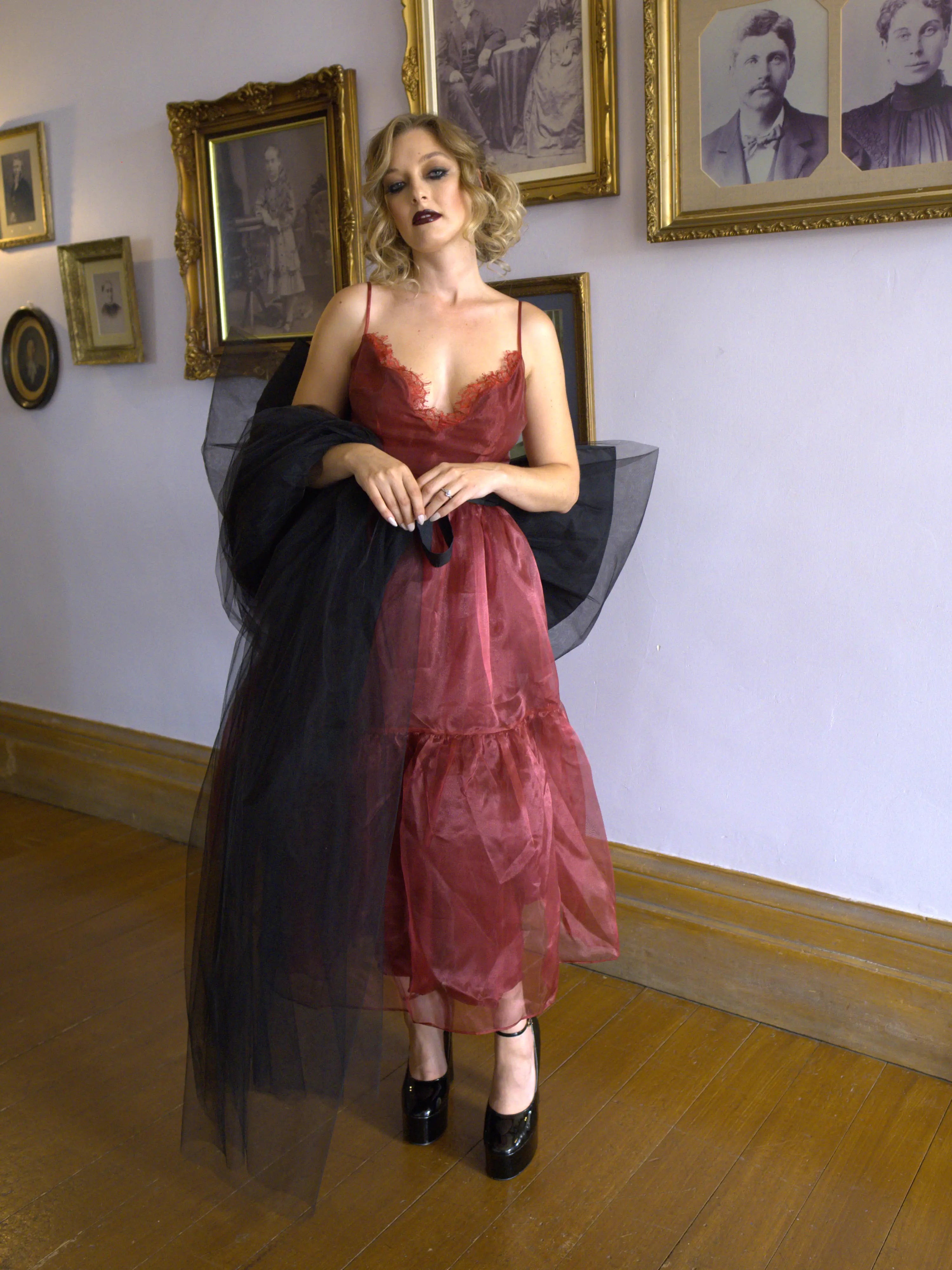 A woman wearing a red organza midi dress with a black tulle giant bow sash in an art gallery setting.