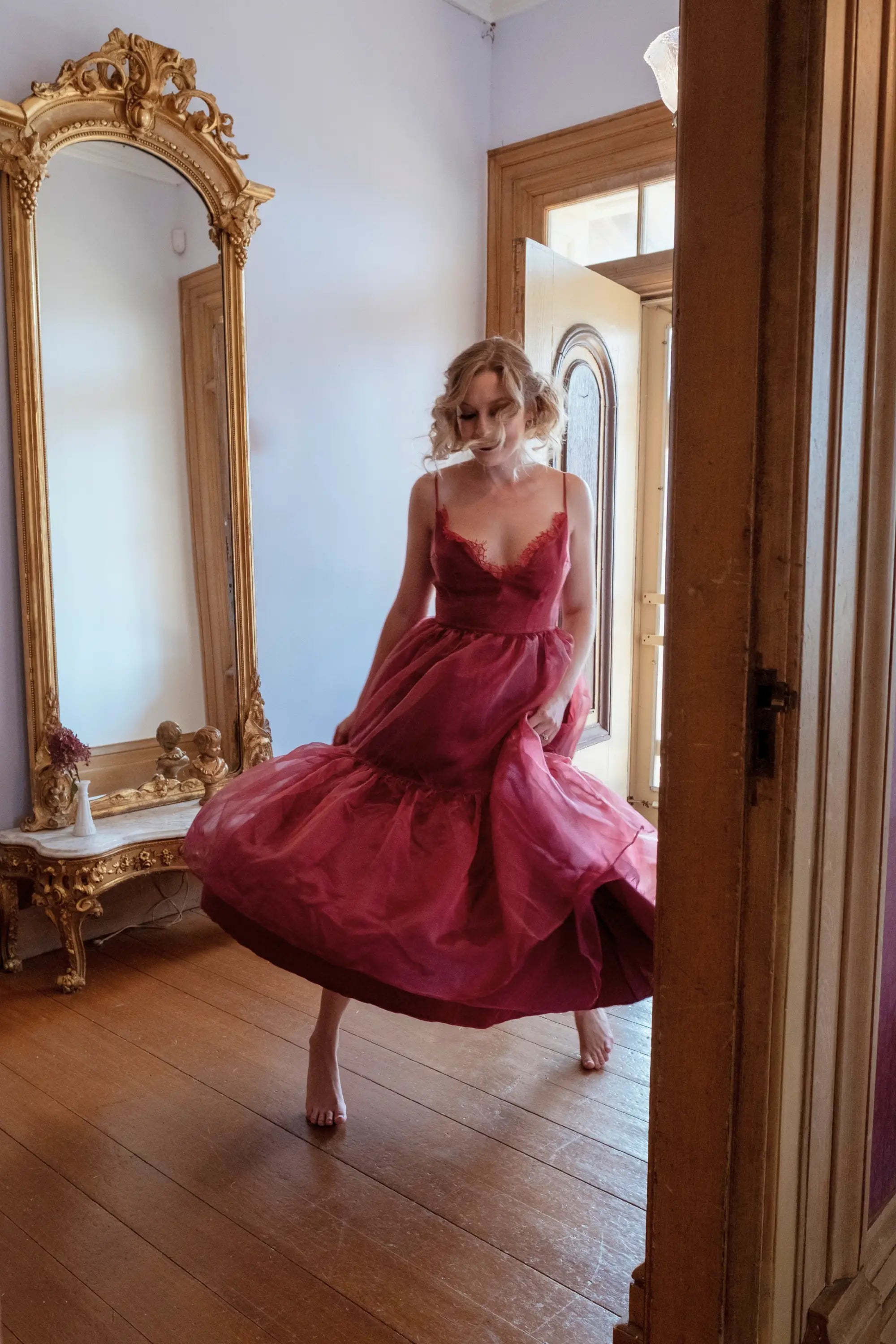 A woman in a red organza midi dress standing in a room with wooden floors and large mirrors.