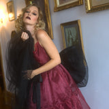 A woman in a red organza midi dress holding a black organza giant waist bow in a room with framed pictures on the wall.