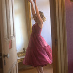 A woman in the Harlow Crimson Organza Midi Dress is standing in front of a large mirror in a room with wooden floors and a gold-framed mirror.