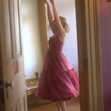A woman in the Harlow Crimson Organza Midi Dress is standing in front of a large mirror in a room with wooden floors and a gold-framed mirror.