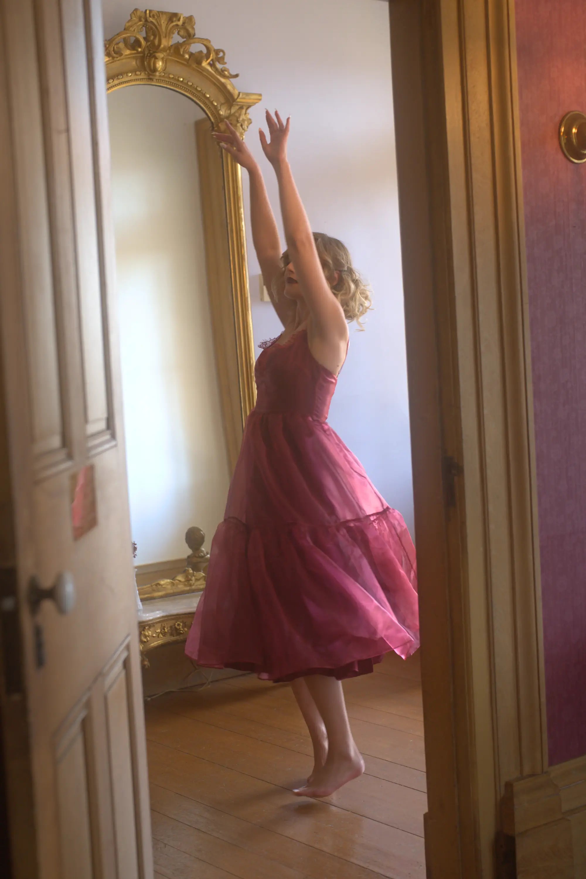 A woman in the Harlow Crimson Organza Midi Dress is standing in front of a large mirror in a room with wooden floors and a gold-framed mirror.
