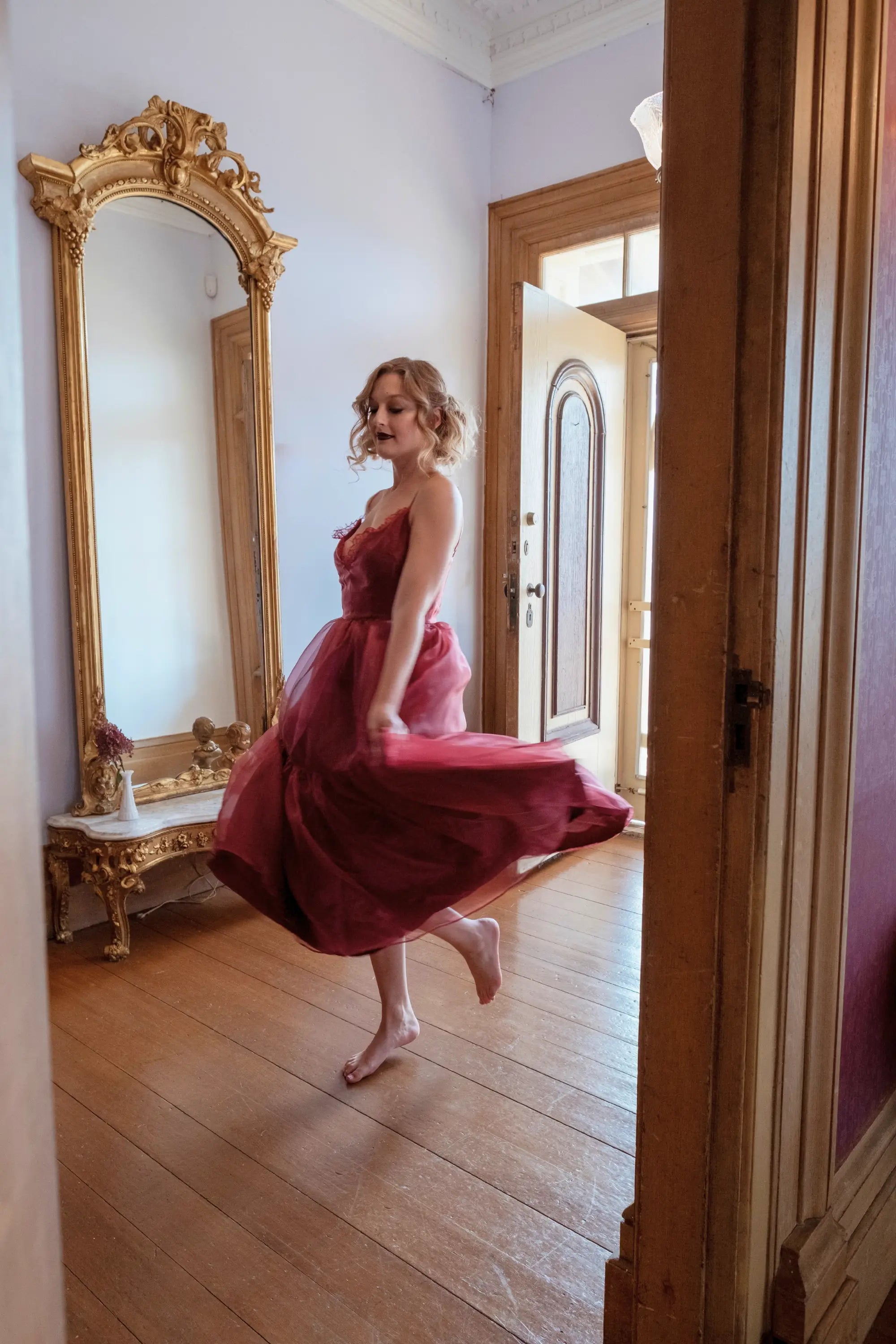 A woman in the Harlow Crimson Organza Midi Dress is standing in a room with wooden floors and an ornate mirror.