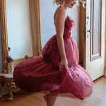 A woman in the Harlow Crimson Organza Midi Dress is standing in a room with a wooden floor and a large mirror.
