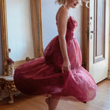 A woman in the Harlow Crimson Organza Midi Dress is standing in a room with a wooden floor and a large mirror.