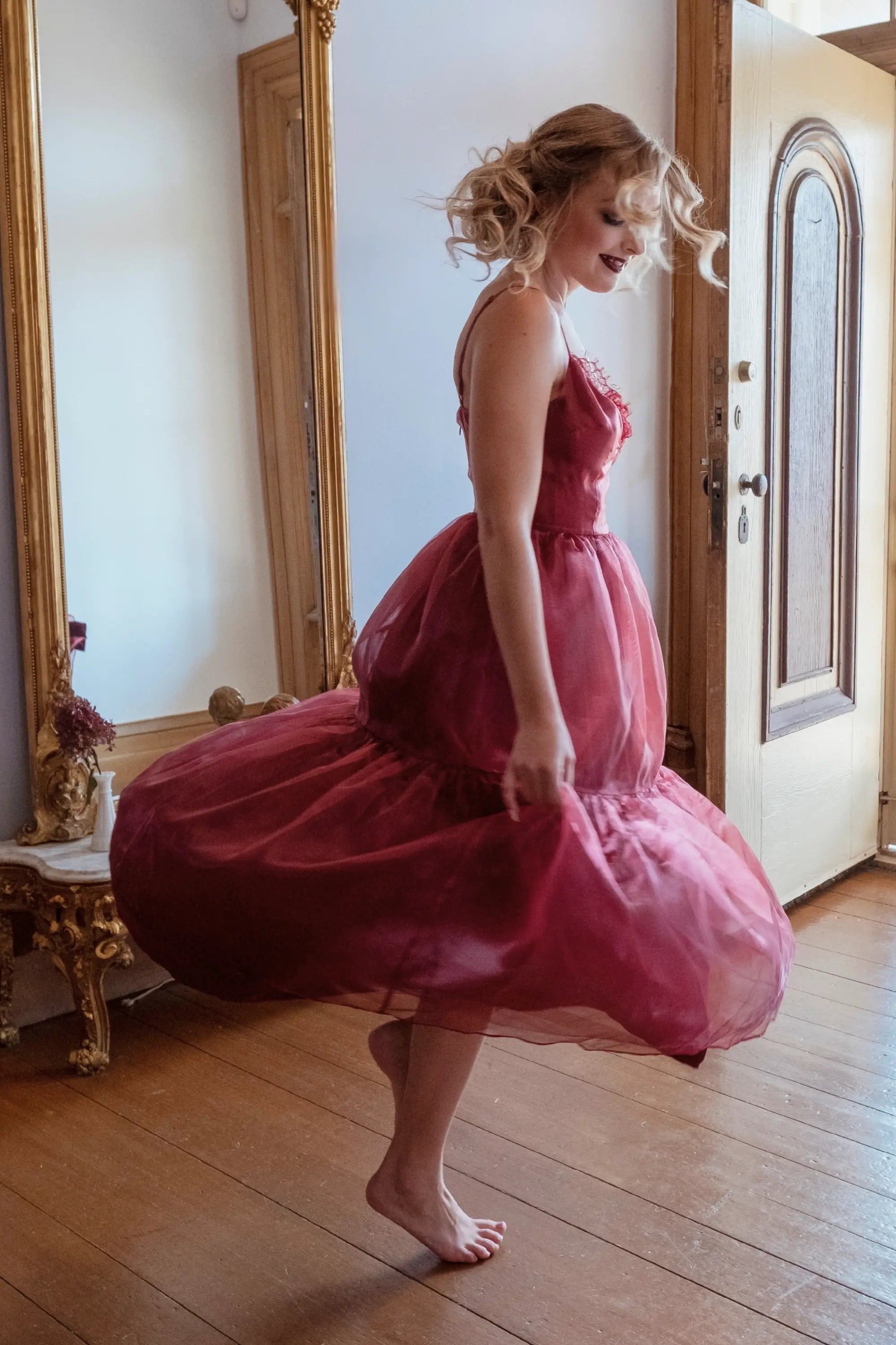 A woman in the Harlow Crimson Organza Midi Dress is standing in a room with a wooden floor and a large mirror.
