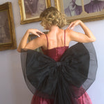 A woman wearing a red organza midi dress with a large black waist bow in front of vintage portraits.