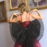 A woman wearing a red organza midi dress with a large black waist bow in front of vintage portraits.