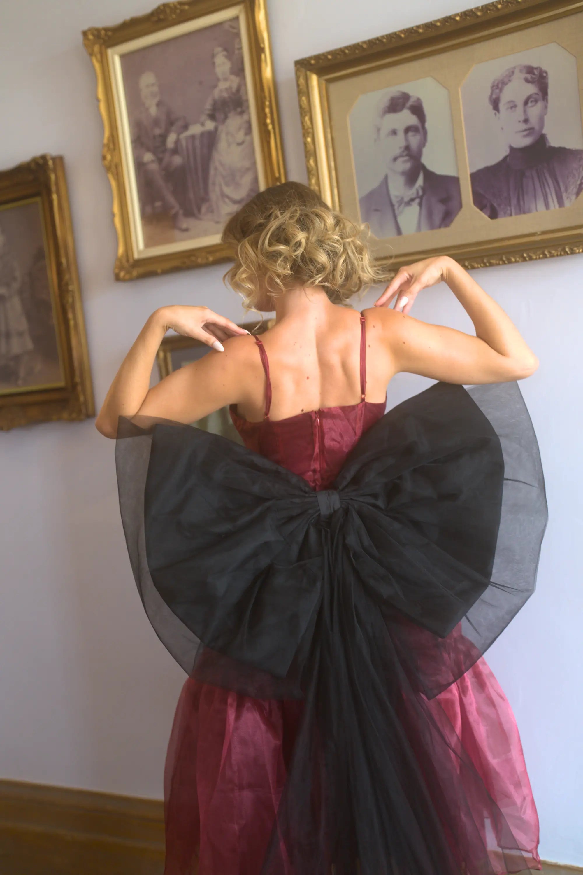A woman wearing a red organza midi dress with a large black waist bow in front of vintage portraits.