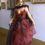 A woman wearing the Harlow crimson red organza midi dress with a black tulle bow sash belt, standing in an art gallery.