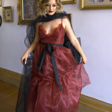 A woman wearing the Harlow crimson red organza midi dress with a black tulle bow sash belt, standing in an art gallery.