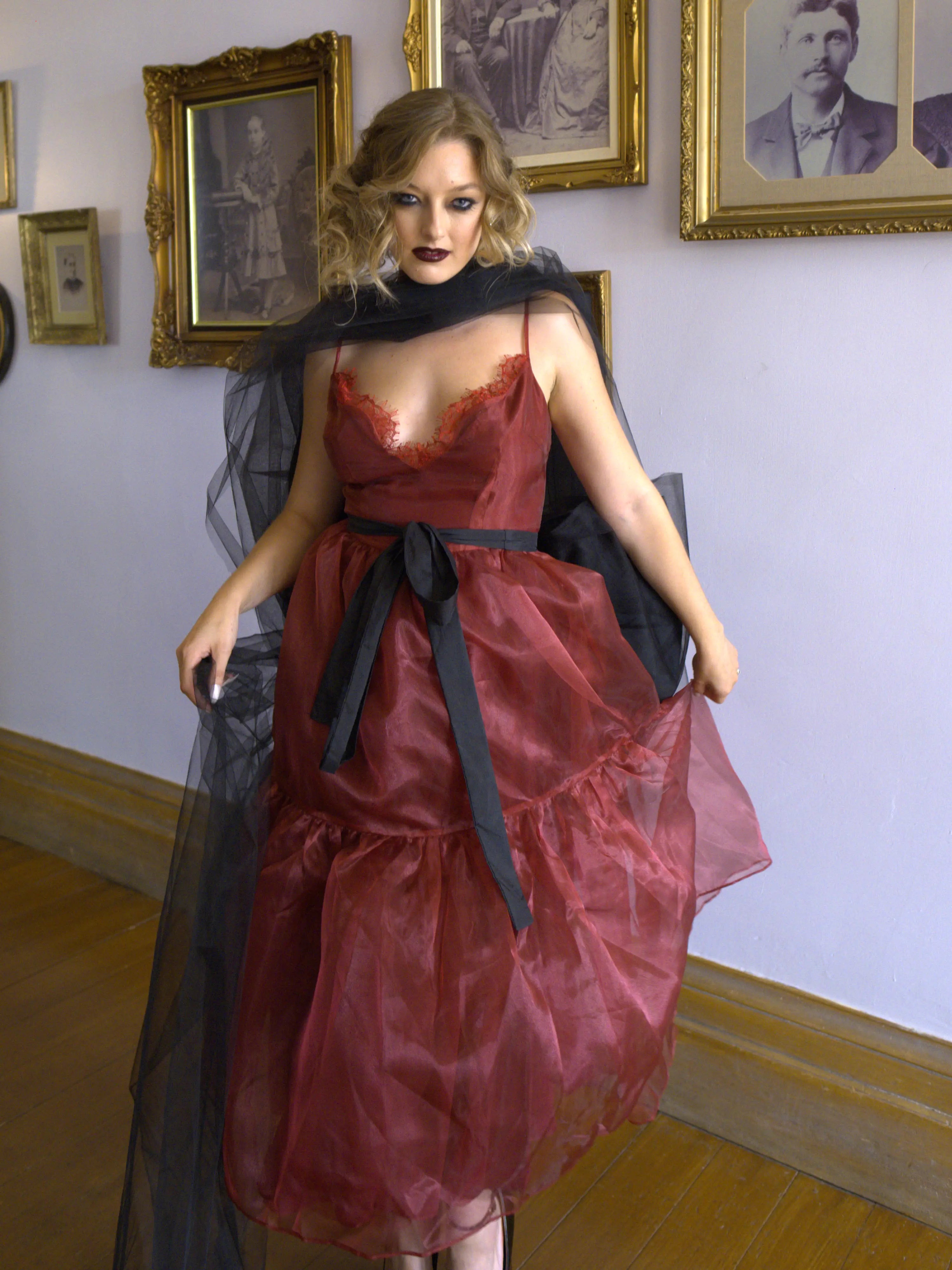 A woman wearing the Harlow crimson red organza midi dress with a black tulle bow sash belt, standing in an art gallery.