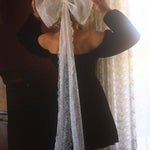 A person wearing a black mini dress with a large white lace hair bow on the back, standing in front of a window with curtains.