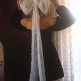 A person wearing a black mini dress with a large white lace hair bow on the back, standing in front of a window with curtains.