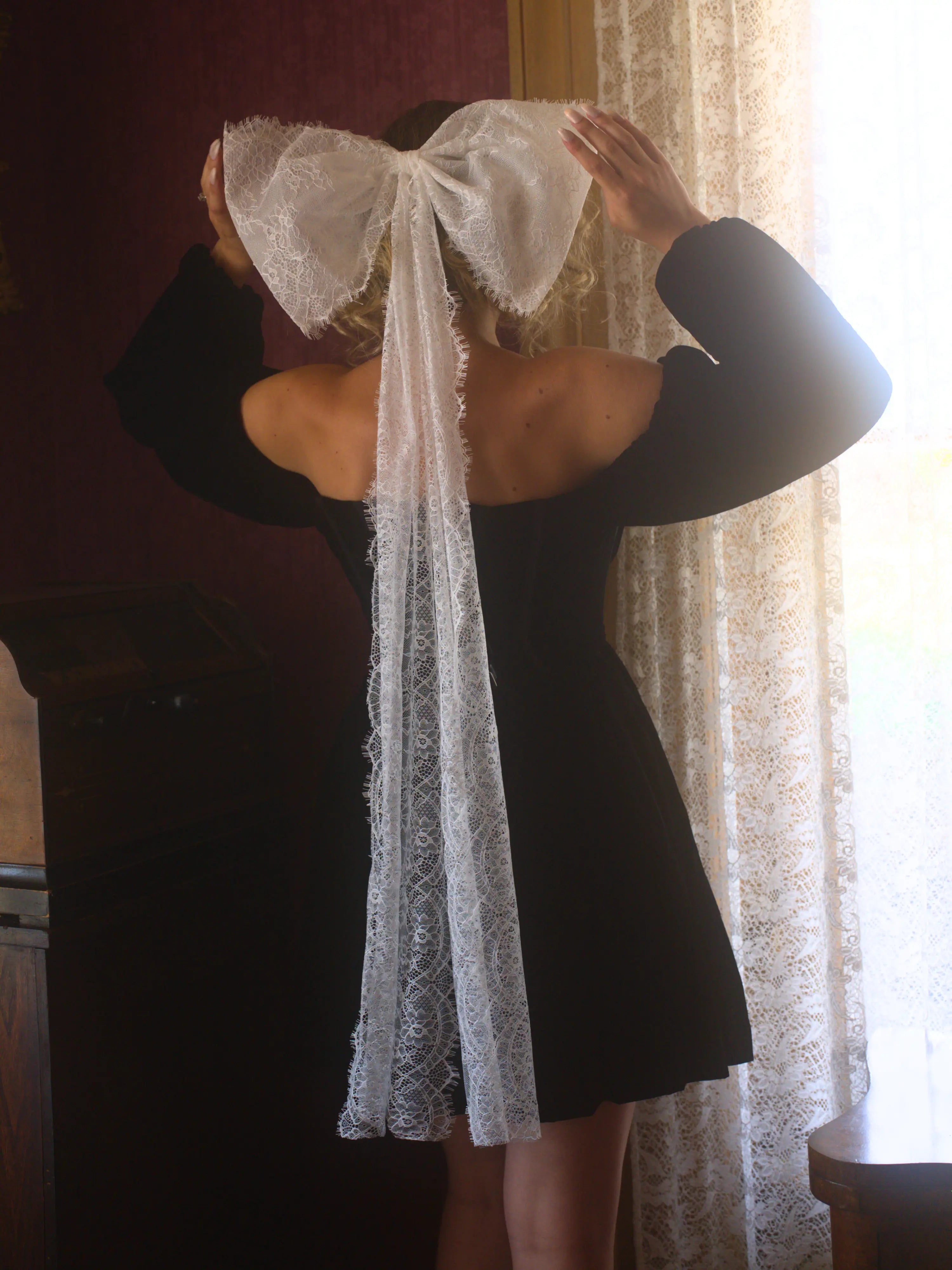 A person wearing a black mini dress with a large white lace hair bow on the back, standing in front of a window with curtains.