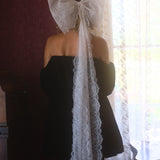A person wearing a black velvet mini dress with a large white lace hair bow on their head, standing in front of a window.