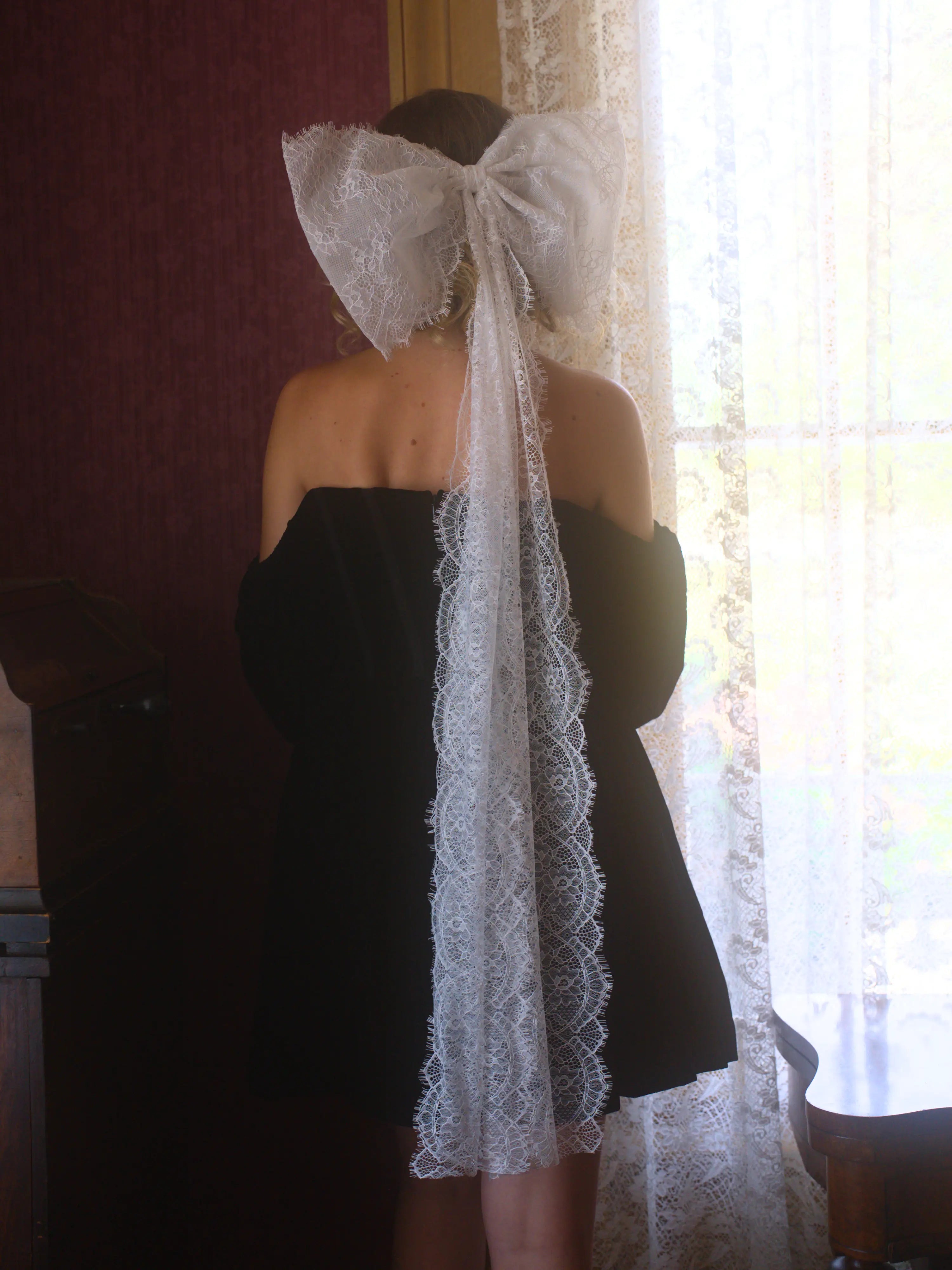 A person wearing a black velvet mini dress with a large white lace hair bow on their head, standing in front of a window.