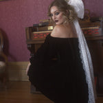 A woman in a black off-shoulder dress with a white lace hair bow in a vintage room.