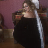 A woman in a black off-shoulder dress with a white lace hair bow in a vintage room.