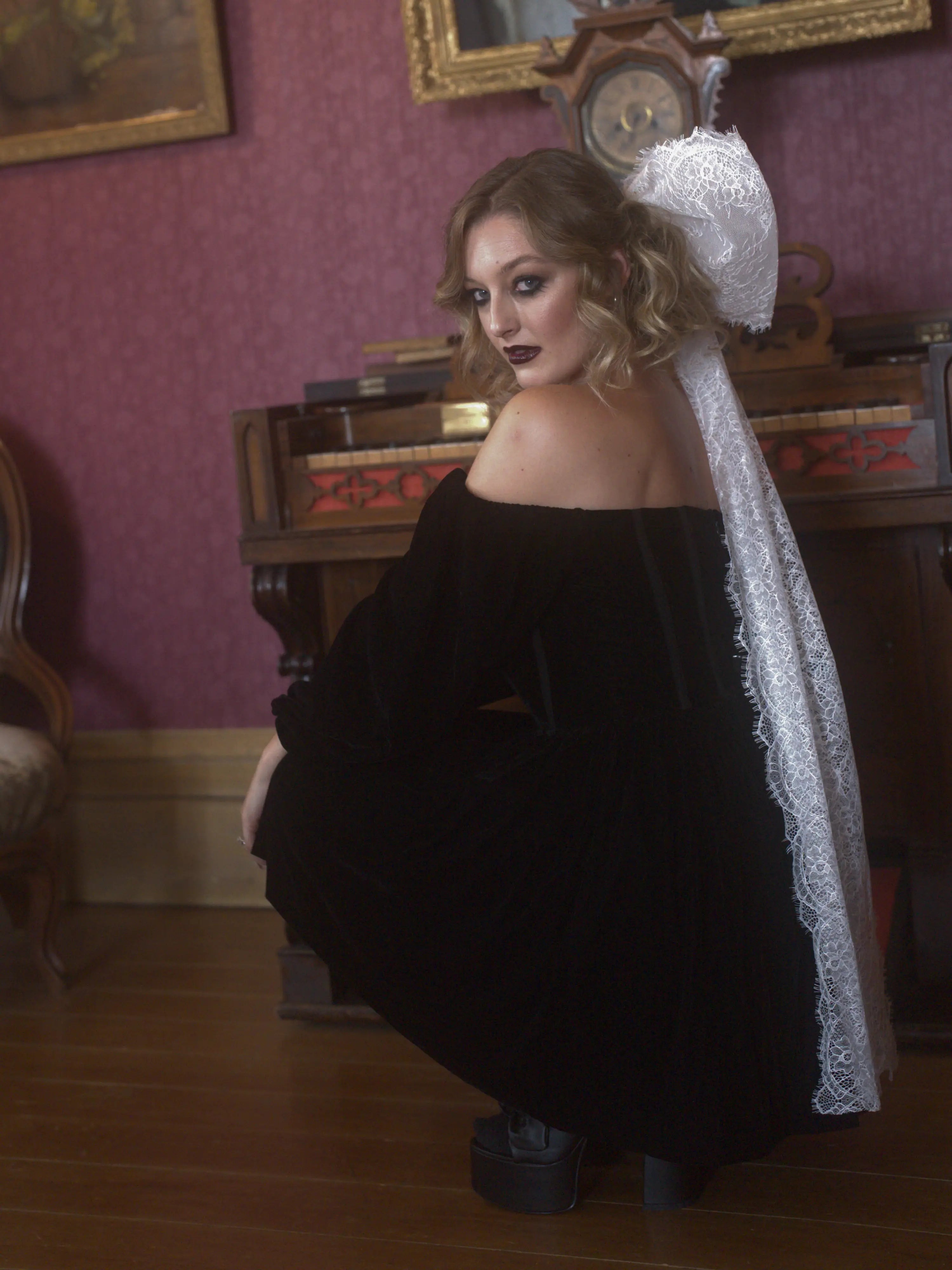 A woman in a black off-shoulder dress with a white lace hair bow in a vintage room.