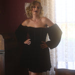 A person wearing a black off-shoulder mini dress with the heirloom lace grand hair bow in a room with curtains and a table.