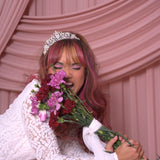 A woman wearing Ivory Sheep's Eloise Lace Mini Dress with a tiara, biting flowers against a pink curtain background.