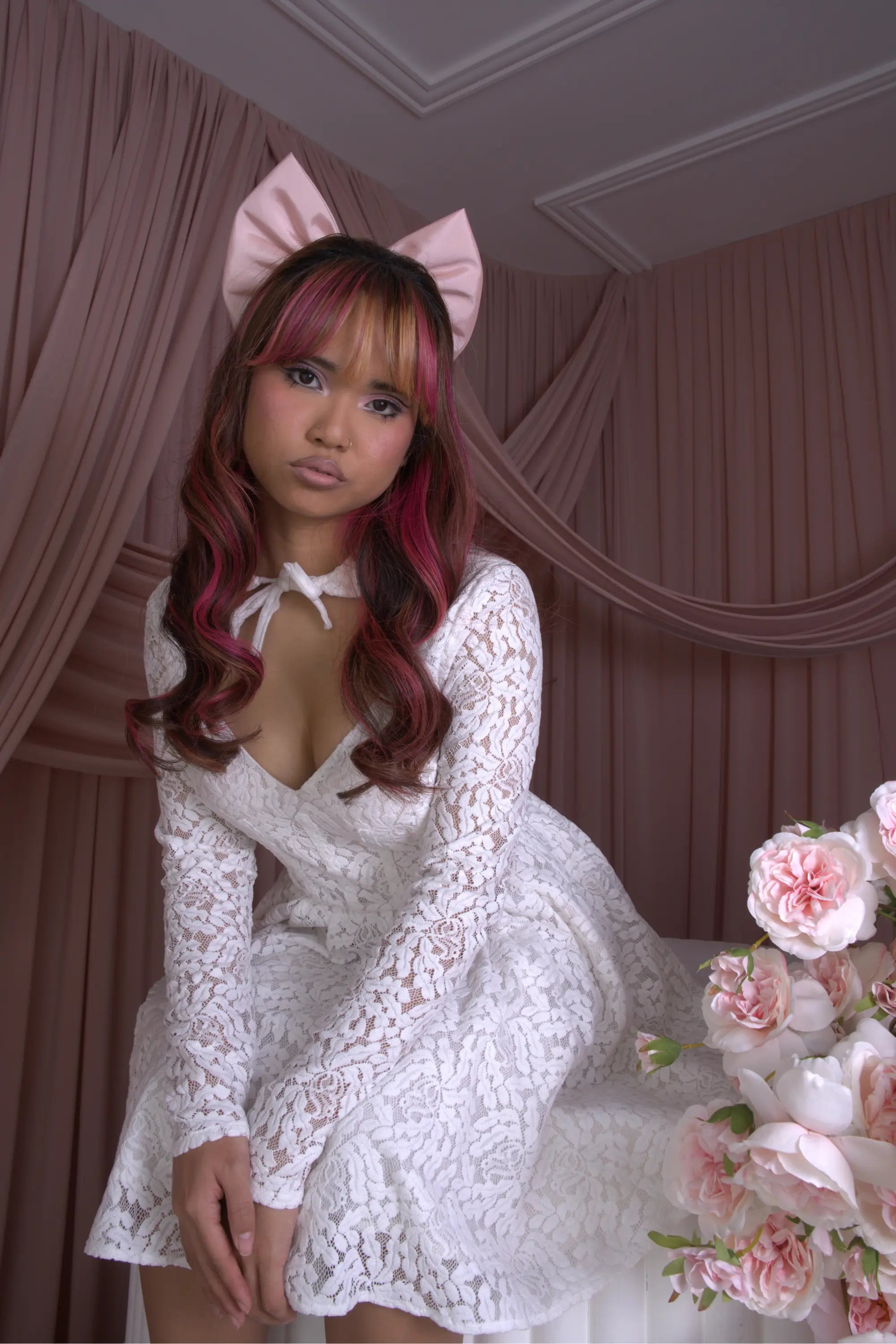 A woman wearing Ivory Sheep's Eloise Mini Lace Dress with a pink hair bow, sitting next to a floral arrangement in a room with pink curtains.